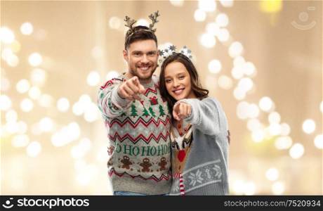christmas, people and holidays concept - happy couple at ugly sweater party pointing at you over festive lights background. happy couple at christmas ugly sweater party