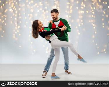 christmas, people and holidays concept - happy couple dancing at ugly sweater party over festive lights background. couple dancing at christmas ugly sweater party