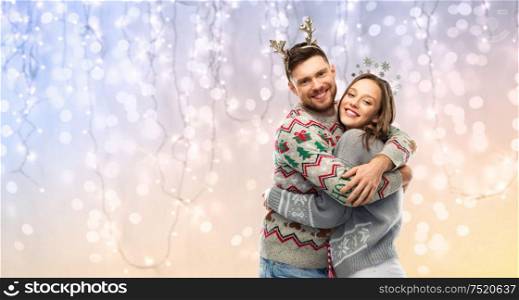 christmas, people and holidays concept - happy couple hugging at ugly sweater party over festive lights background. happy couple at christmas ugly sweater party