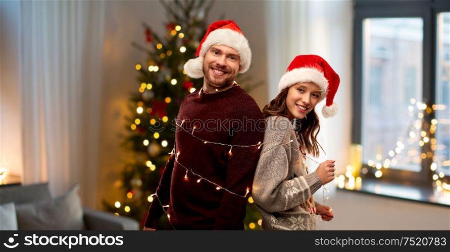 christmas, people and holidays concept - happy couple in santa hats tied by electric garland string over home background. couple in santa hats tied by christmas garland