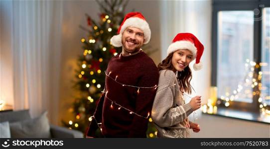 christmas, people and holidays concept - happy couple in santa hats tied by electric garland string over home background. couple in santa hats tied by christmas garland