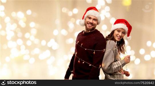 christmas, people and holidays concept - happy couple in santa hats tied by electric garland string over festive lights background. couple in santa hats tied by christmas garland