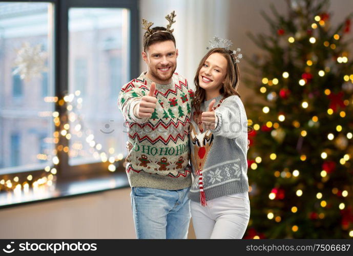 christmas, people and holidays concept - happy couple showing thumbs up at ugly sweater party over home room background. couple in christmas ugly sweaters shows thumbs up