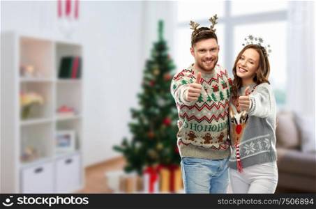 christmas, people and holidays concept - happy couple showing thumbs up at ugly sweater party over home room background. couple in christmas ugly sweaters shows thumbs up