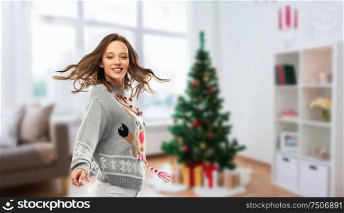 christmas, people and holidays concept - happy young woman in jumper with reindeer pattern dancing at ugly sweater party over home room background. woman dancing at christmas ugly sweater party