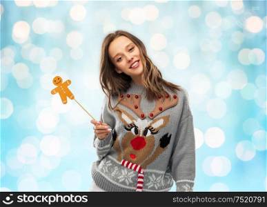 christmas, people and holidays concept - happy young woman in jumper with reindeer pattern holding gingerbread accessory at ugly sweater party over festive lights on blue background. woman in christmas sweater with party accessory