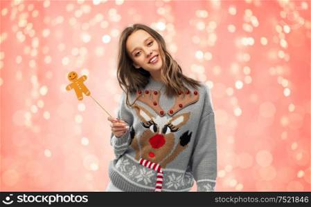 christmas, people and holidays concept - happy young woman in jumper with reindeer pattern holding gingerbread accessory at ugly sweater party over festive lights on pink coral background. woman in christmas sweater with party accessory