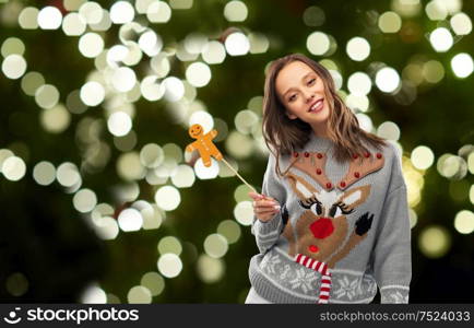 christmas, people and holidays concept - happy young woman in jumper with reindeer pattern holding gingerbread accessory at ugly sweater party over festive lights on dark green background. woman in christmas sweater with reindeer pattern