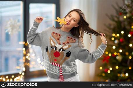 christmas, people and holidays concept - happy young woman wearing jumper with reindeer pattern and biting gingerbread party accessory at ugly sweater party over home room background. woman in christmas sweater with reindeer pattern