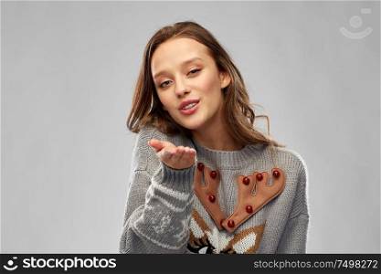 christmas, people and holidays concept - happy young woman wearing ugly sweater with reindeer pattern sending air kiss over grey background. woman in ugly christmas sweater sending air kiss