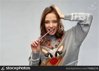 christmas, people and holidays concept - happy young woman wearing ugly sweater with reindeer pattern eating candy cane. woman in christmas sweater eating candy cane