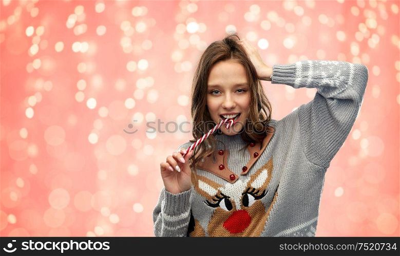 christmas, people and holidays concept - happy young woman wearing ugly sweater with reindeer pattern biting candy cane over festive lights on pink coral background. woman in christmas sweater biting candy cane