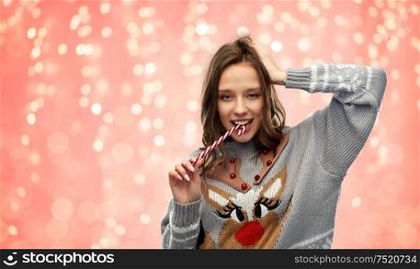 christmas, people and holidays concept - happy young woman wearing ugly sweater with reindeer pattern biting candy cane over festive lights on pink coral background. woman in christmas sweater biting candy cane