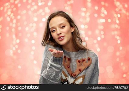 christmas, people and holidays concept - happy young woman wearing ugly sweater with reindeer pattern sending air kiss over festive lights on pink coral background. woman in ugly christmas sweater sending air kiss