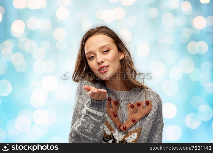 christmas, people and holidays concept - happy young woman wearing ugly sweater with reindeer pattern sending air kiss over festive lights on blue background. woman in ugly christmas sweater sending air kiss