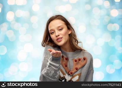 christmas, people and holidays concept - happy young woman wearing ugly sweater with reindeer pattern sending air kiss over festive lights on blue background. woman in ugly christmas sweater sending air kiss