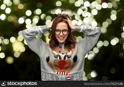 christmas, people and holidays concept - happy young woman wearing ugly sweater with reindeer pattern over festive lights on dark green background. woman in christmas sweater with reindeer pattern