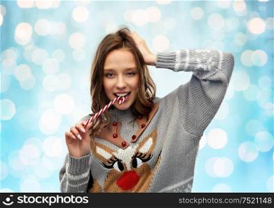 christmas, people and holidays concept - happy young woman wearing ugly sweater with reindeer pattern biting candy cane over festive lights on blue background. woman in christmas sweater biting candy cane