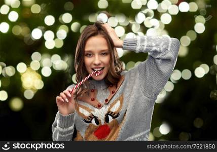 christmas, people and holidays concept - happy young woman wearing ugly sweater with reindeer pattern eating candy cane over festive lights on dark green background. woman in christmas sweater eating candy cane