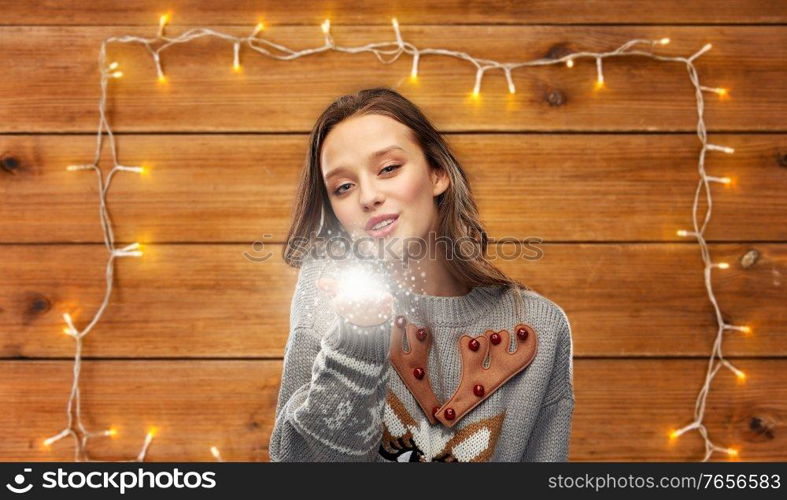 christmas, people and holidays concept - happy young woman wearing ugly sweater with reindeer pattern with magical fairy dust over garland lights on wooden background. woman in ugly christmas sweater with magical dust