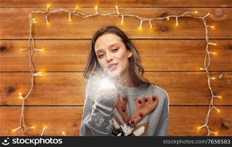 christmas, people and holidays concept - happy young woman wearing ugly sweater with reindeer pattern with magical fairy dust over garland lights on wooden background. woman in ugly christmas sweater with magical dust