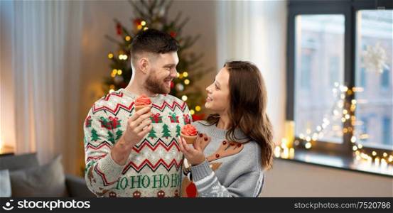 christmas, people and holidays concept - portrait of happy couple at ugly sweater party with cupcakes over home background. couple with cupcakes in ugly christmas sweaters