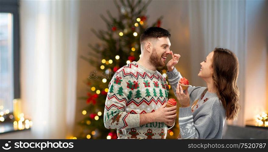 christmas, people and holidays concept - portrait of happy couple at ugly sweater party with cupcakes over home background. couple with cupcakes in ugly christmas sweaters