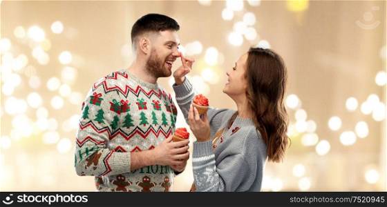 christmas, people and holidays concept - portrait of happy couple at ugly sweater party with cupcakes over festive lights background. couple with cupcakes in ugly christmas sweaters