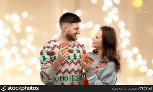 christmas, people and holidays concept - portrait of happy couple at ugly sweater party with cupcakes over festive lights background. couple with cupcakes in ugly christmas sweaters