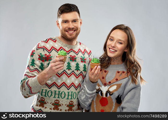 christmas, people and holidays concept - portrait of happy couple at ugly sweater party with cupcakes. couple with cupcakes in ugly christmas sweaters