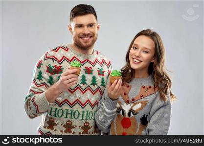 christmas, people and holidays concept - portrait of happy couple at ugly sweater party with cupcakes. couple with cupcakes in ugly christmas sweaters