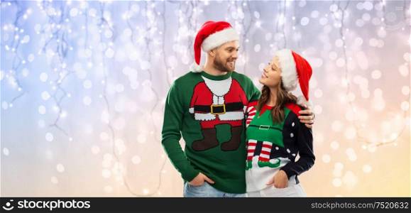 christmas, people and holidays concept - portrait of happy couple in santa hats at ugly sweater party over festive lights background. happy couple in christmas sweaters and santa hats