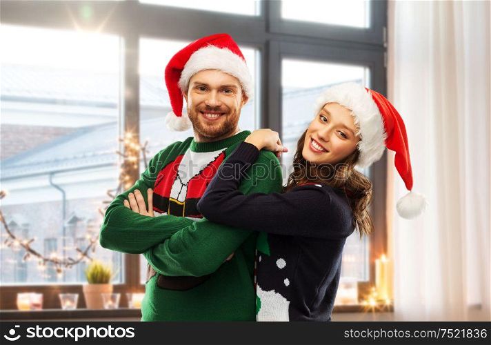 christmas, people and holidays concept - portrait of happy couple in santa hats at ugly sweater party over home room background. happy couple in christmas sweaters and santa hats
