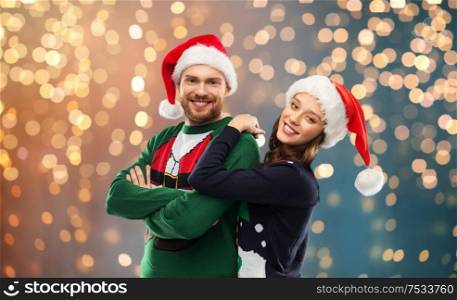 christmas, people and holidays concept - portrait of happy couple in santa hats at ugly sweater party over festive lights background. happy couple in christmas sweaters and santa hats