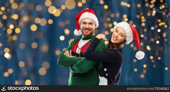 christmas, people and holidays concept - portrait of happy couple in santa hats at ugly sweater party over festive lights on dark night background. happy couple in christmas sweaters and santa hats