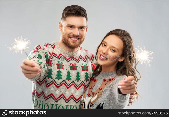 christmas, people and holidays concept - portrait of happy couple with sparklers at ugly sweater party. happy couple in christmas sweaters with sparklers