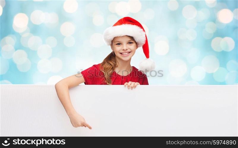 christmas, people, children and advertisement concept - happy girl child in santa helper hat pointing finger on blank white board over blue holidays lights background