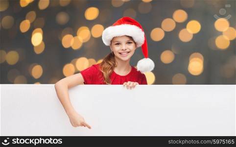 christmas, people, children and advertisement concept - happy girl child in santa helper hat pointing finger on blank white board over golden lights background