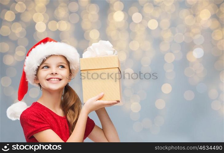 christmas, people, children and holidays concept - smiling girl in santa helper hat with gift box over lights background