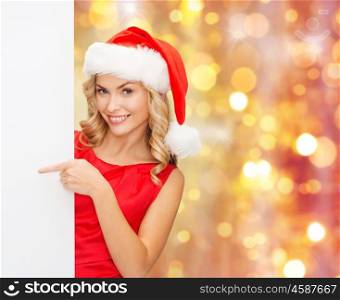 christmas, people, holidays and advertisement concept - happy woman in santa helper hat pointing finger to blank white board over lights background