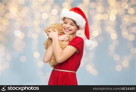 christmas, people, holidays and childhood concept - smiling girl in santa helper hat hugging teddy bear over lights background