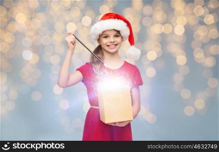 christmas, people, holidays and magic concept - smiling girl in santa helper hat with gift box and magic wand over lights background. girl in santa hat with gift box and magic wand