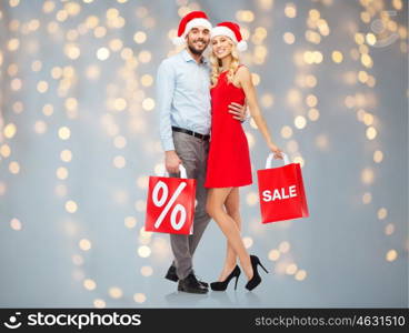 christmas, people, sale, discount and holidays concept - happy couple in santa hats hugging with red shopping bags over lights background