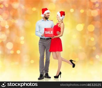 christmas, people, sale, discount and holidays concept - happy couple in santa hats with red sale sign over lights background