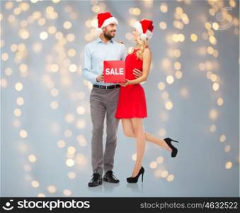christmas, people, sale, discount and holidays concept - happy couple in santa hats with red sale sign over lights background