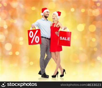 christmas, people, sale, discount and holidays concept - happy couple in santa hats hugging with red shopping bags over lights background