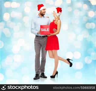 christmas, people, sale, discount and holidays concept - happy couple in santa hats with red sale sign over blue lights background