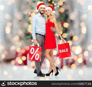 christmas, people, sale, discount and holidays concept - happy couple in santa hats hugging with red shopping bags over christmas tree lights background