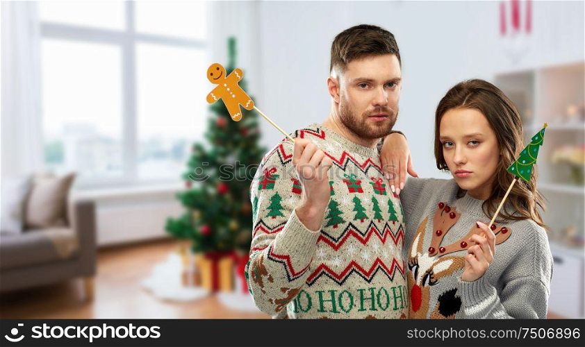 christmas, photo booth and holidays concept - gloomy couple in ugly sweaters posing with party props over home room background. couple with christmas party props in ugly sweaters