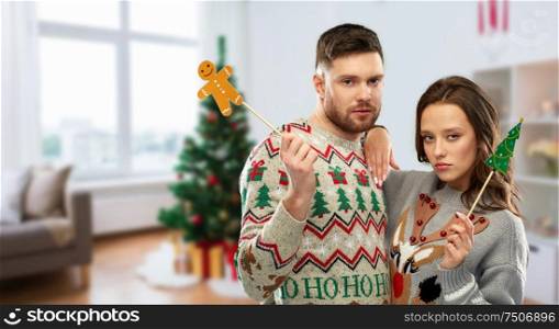 christmas, photo booth and holidays concept - gloomy couple in ugly sweaters posing with party props over home room background. couple with christmas party props in ugly sweaters